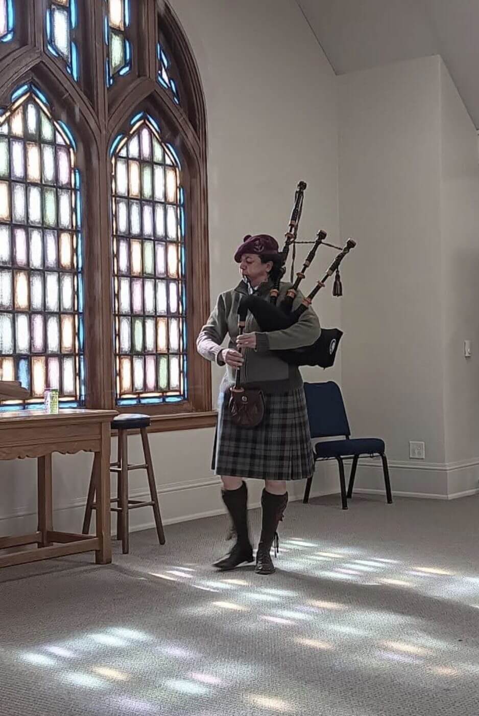 A Rose & Thistle piper in tartan kilt and beret playing the Great Highland bagpipes beside a stained-glass window